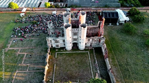 aerial drone shot mid angle flying away and panning up tracking ancient old English Titchfield abbey ruins with large crowd of people in countryside in summer at sunset