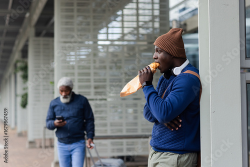 African american male leaning on pillar eating sandwich with headphones under canopy