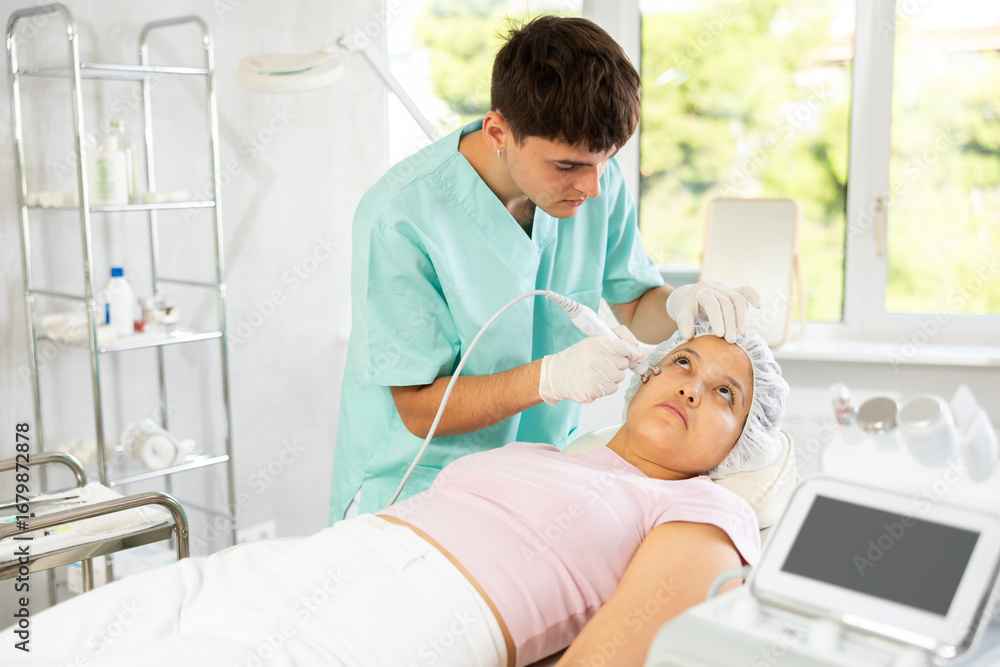 Obraz premium Young male cosmetologist performs facial microcurrent procedure with machine to young female client