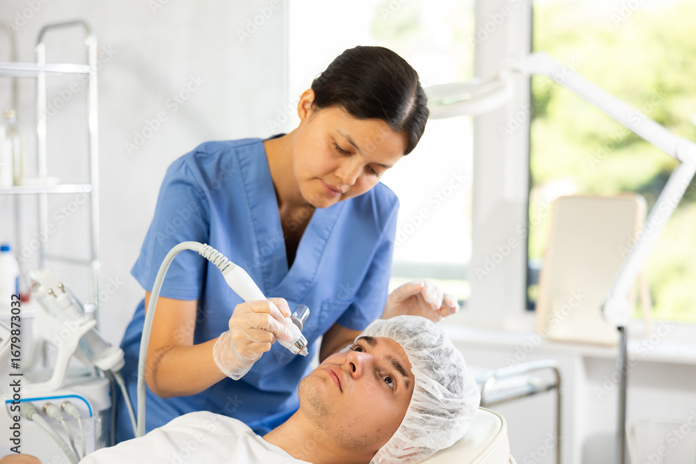 Fototapeta premium Young female cosmetologist performs hardware facial moisturizing procedure to young male patient