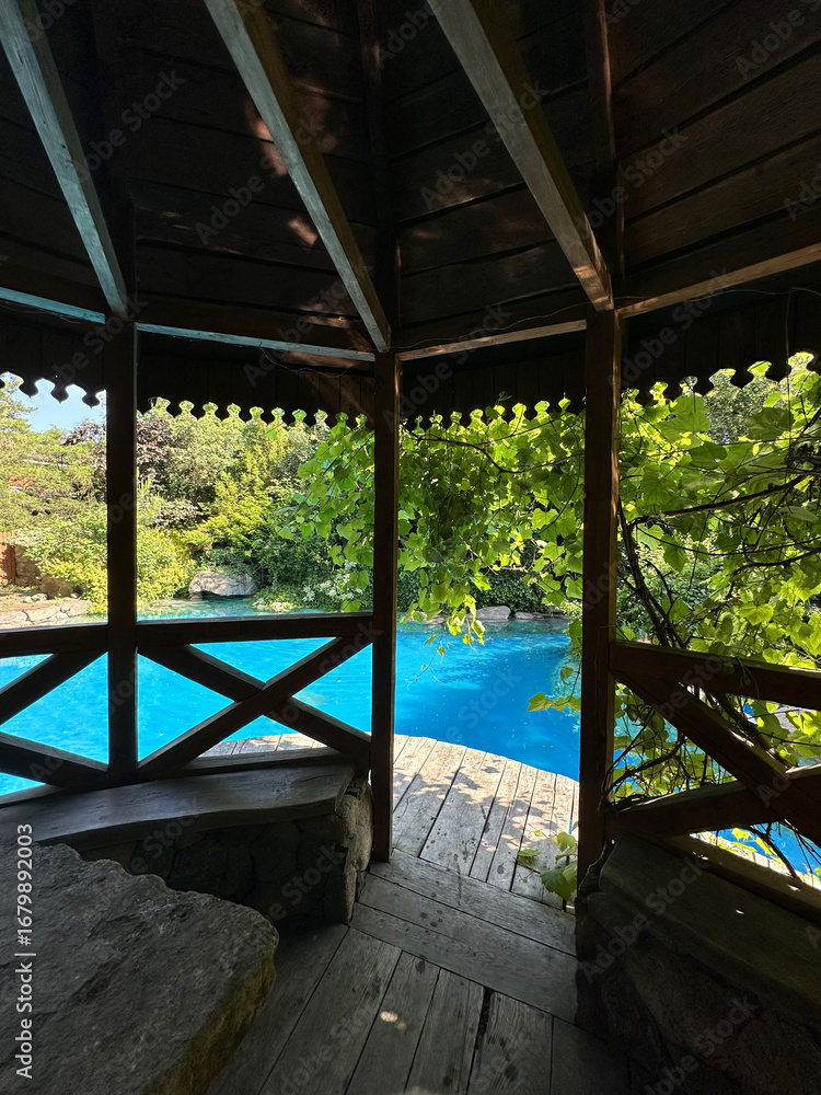 Fototapeta premium Cozy wooden gazebo surrounded by lush greenery, overlooking a serene blue swimming pool, creating a tranquil outdoor retreat for relaxation and leisure