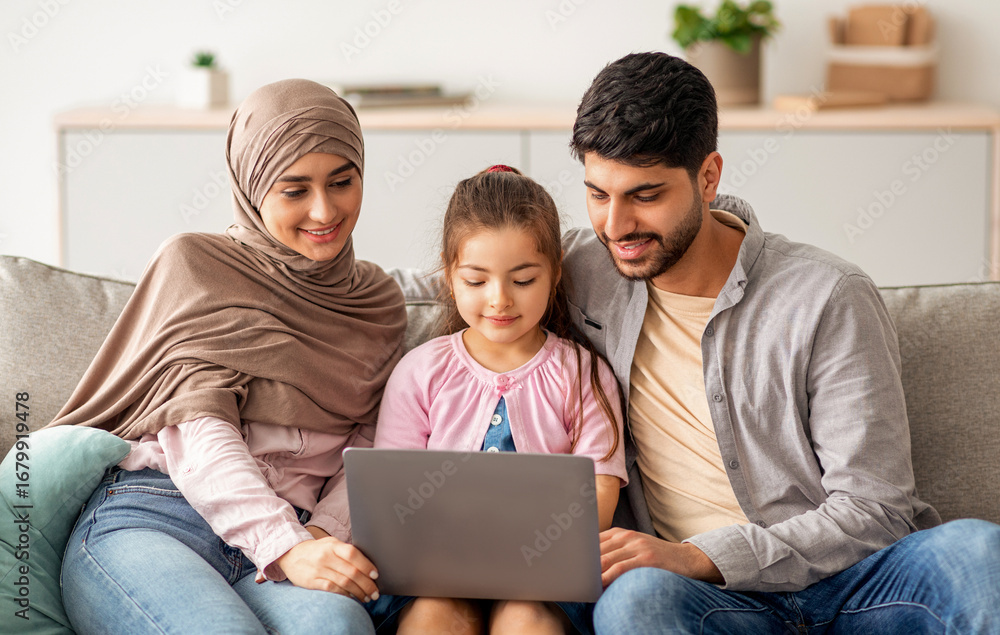 Fototapeta premium Cheerful muslim family having fun with laptop, surfing internet, playing games or having video chat, sitting together on sofa. Happy arab man, child and woman in hijab resting at home