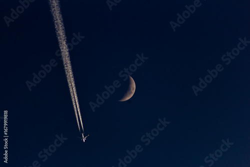 A plane in the night sky with the moon and contrail.