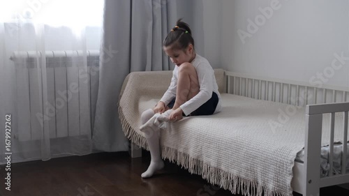 Side view of a young girl sitting on a bed and putting on tights in a bedroom at home.
