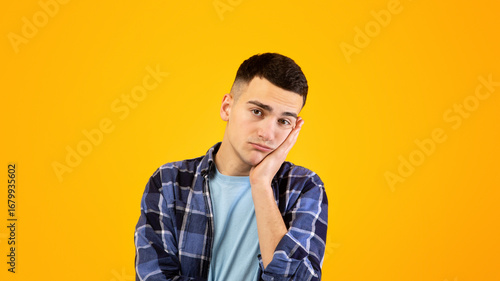 Portrait of young guy feeling bored, leaning on his hand, having dull day on orange studio background. Funky Caucasian man being annoyed or disappointed, showing lack of interest in something