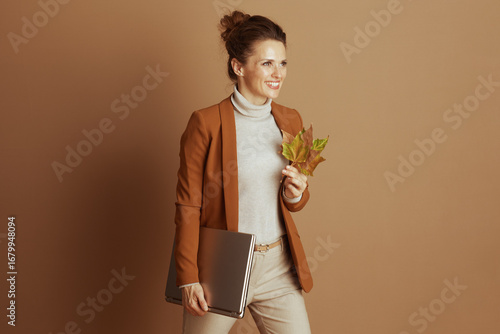 A smiling professional woman in a brown blazer and beige turtleneck confidently holds a closed silver laptop and autumn leaves. She represents a cheerful, modern, seasonally aware business approach.