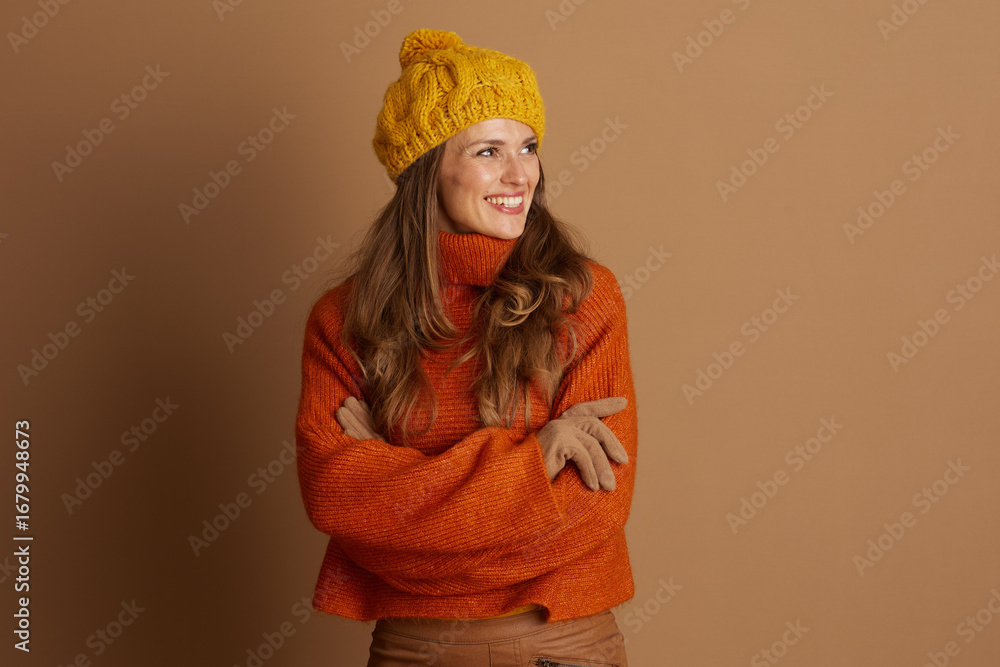 Fototapeta premium A smiling woman in a yellow pom-pom beanie and orange turtleneck, with arms crossed, looks to the side. Against a plain brown background, she presents a cheerful, stylish autumnal or winter look.