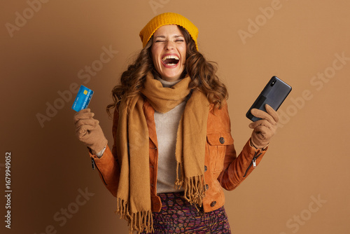 A joyfully laughing woman in autumn fashion holds a credit card and smartphone, embodying the excitement and convenience of digital shopping and financial transactions.
