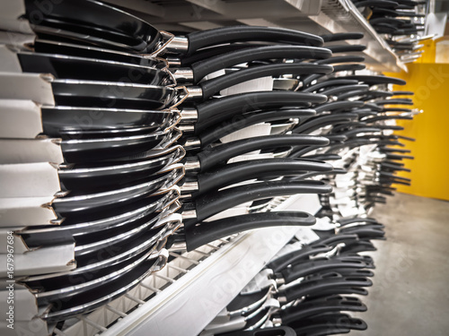 Rows of nonstick pancake pans with long black handles arranged on supermarket shelves. The perspective shows a cookware display, highlighting Teflon coated skillets and consumer choice in the kitchen 