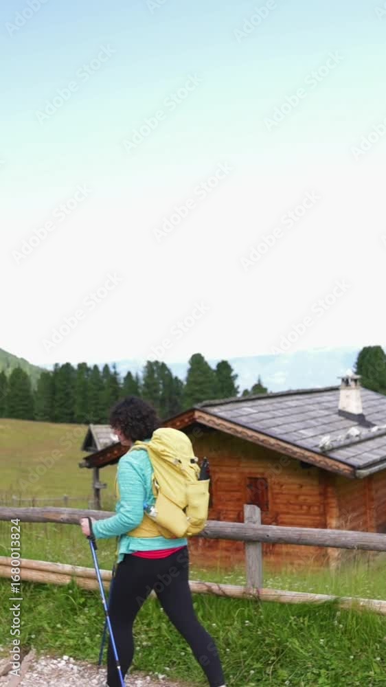 Vertical screen Female hiker on alpine trail near rustic cabins and forest in Italy Hiker with yellow backpack and trekking poles walking past wooden mountain cabins in the Dolomites, Italy Vertical screen Female hiker on alpine trail near rustic cabins and forest in Italy Hiker with yellow backpack and trekking poles walking past wooden mountain cabins in the Dolomites, Italy