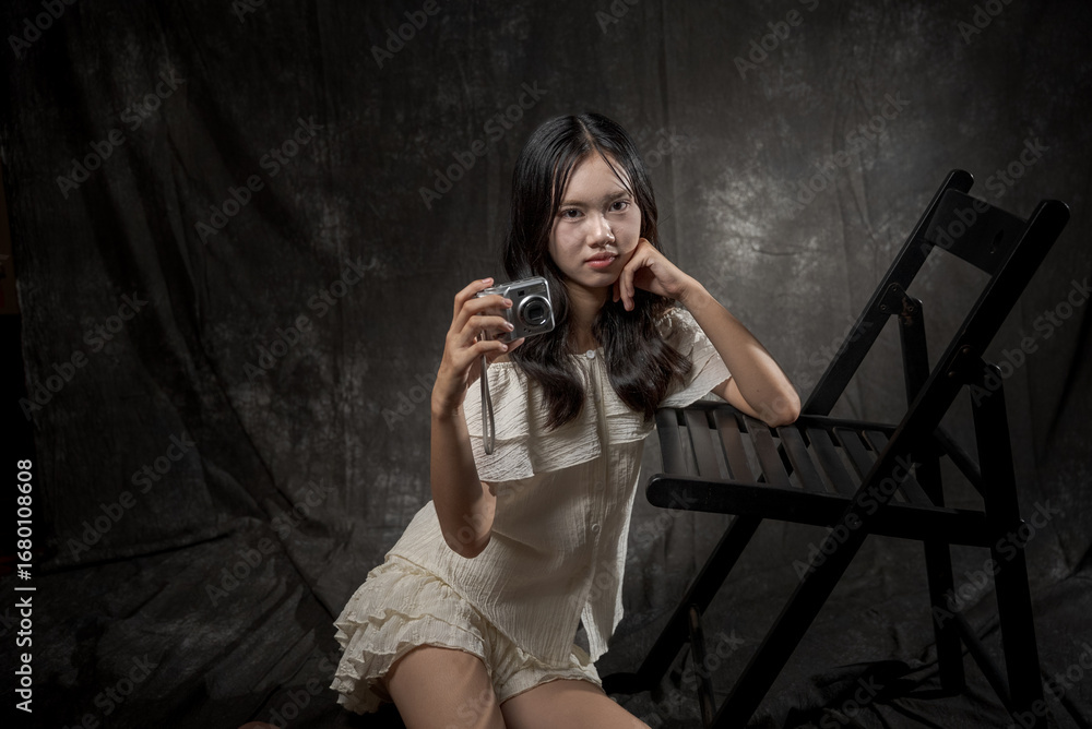 Obraz premium Young Asian woman standing and smiling in studio, wearing off-shoulder white blouse and layered skirt, posing gracefully with one hand on chest and one leg raised, dark background.
