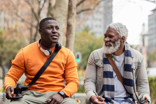 African american male friends sitting on park bench in city square using smartphone with headphones