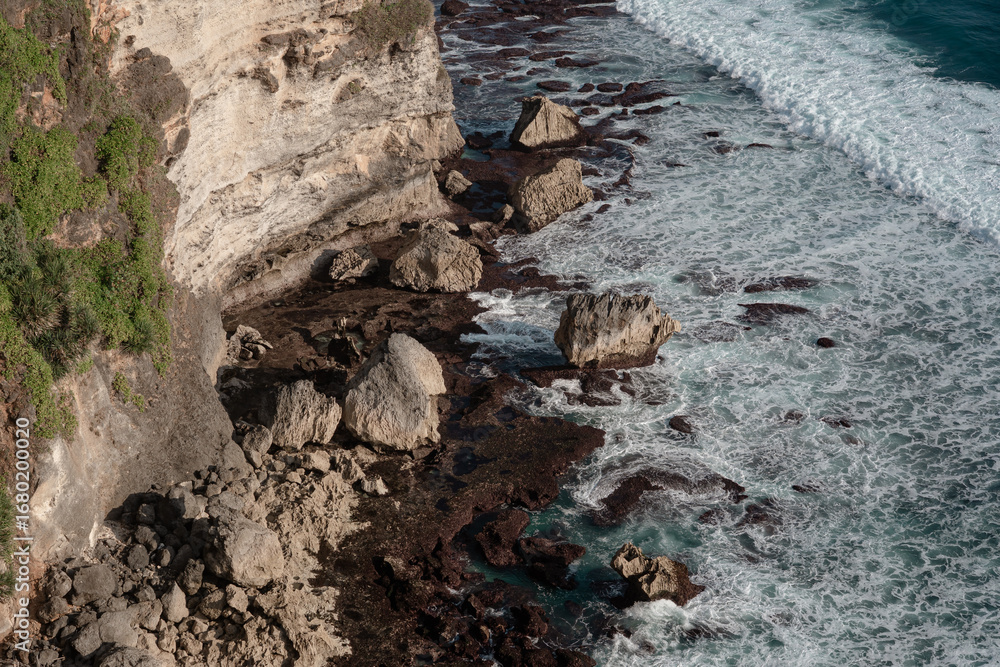 Obraz premium Dramatic view of the rocky base of a large cliff with powerful ocean waves and surf. A wild and untamed coastal landscape in Bali. 