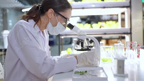A female scientist wearing lab coat and mask conducts scientific experiments with microscope and test tubes in high tech lab.