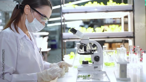 A female scientist wearing lab coat and mask conducts scientific experiments with microscope and test tubes in high tech lab.