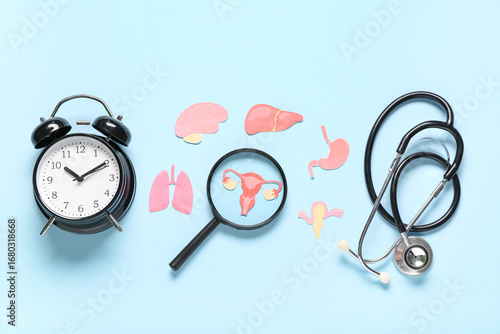 Alarm clock with stethoscope, paper woman's organs and magnifier on blue back...