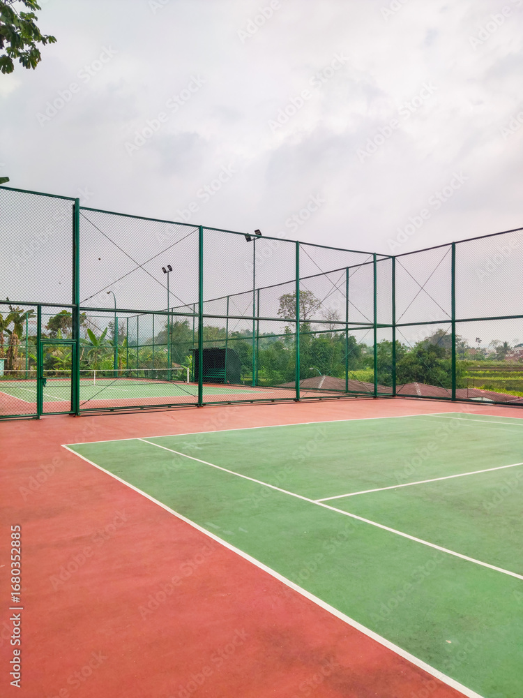 Fototapeta premium Empty tennis court with green and red surface, surrounded by tall wire fencing under an overcast sky, creating a quiet atmosphere for practice, competition, or leisure play in a peaceful outdoor setti