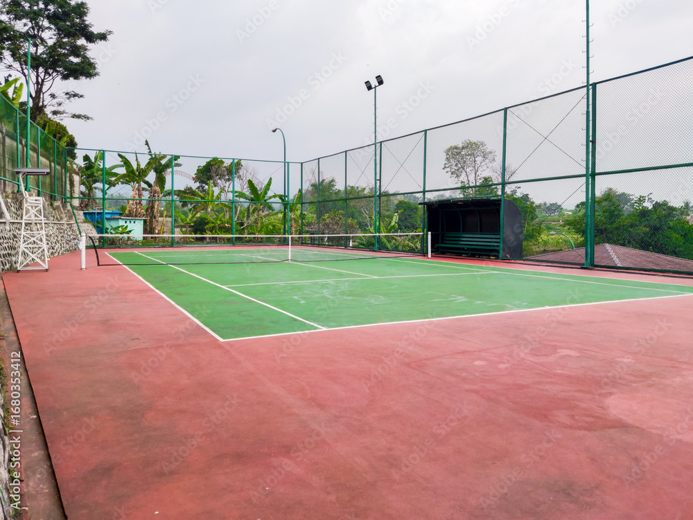 Fototapeta premium Empty tennis court with green and red surface, surrounded by tall wire fencing under an overcast sky, creating a quiet atmosphere for practice, competition, or leisure play in a peaceful outdoor setti