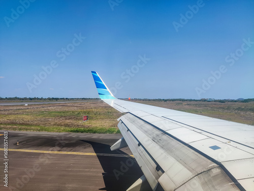 Airplane wing view during taxi on runway, clear blue sky, travel concept, aviation industry, airport ground, jet engine detail, aircraft design, flight preparation, and passenger perspective.