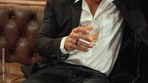 Men's hands hold a glass of whiskey while sitting on a leather sofa.