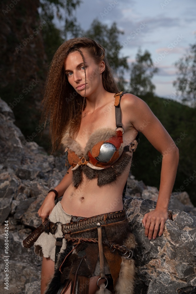 Obraz premium Warrior woman poses amidst rocky terrain, adorned with tribal-inspired armor and weaponry.