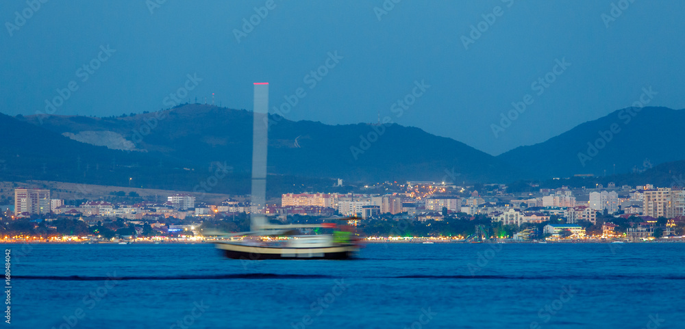 Fototapeta premium Gelecik resort in evening twilight. Pleasure vessel moving along Gelendzhik Bay. City lights and Caucasus Mountains in background.