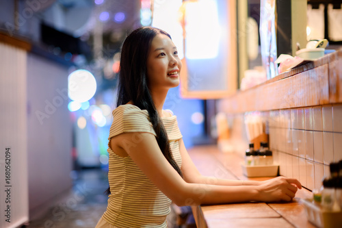 asian woman sitting at the bar counter in a japanese ramen restaurant, holding chopsticks with a smile, warm lights and cozy dining atmosphere.
