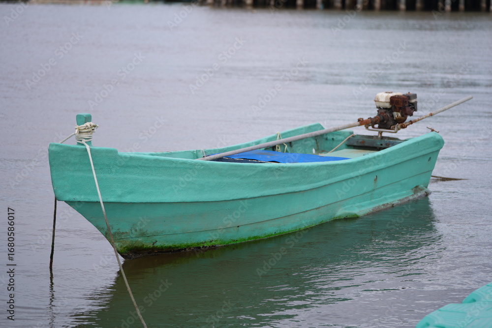 Naklejka premium A light green long-tail boat with an old engine floats calmly on the water, secured with a rope to the dock.