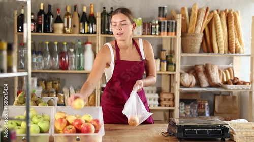 Young saleswoman in uniform weighing apples on scales in grocery store