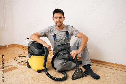 Portrait of professional cleaning service employee in overall sitting on floor next to washing vacuum cleaner, ready for thorough residential floor cleaning and sanitizing service, looking at camera