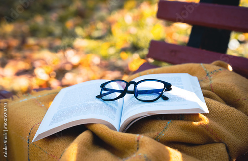 Open book with eyeglasses placed on a warm blanket outdoors on a bench in autumn park. Cozy fall atmosphere, reading, studying, and relaxation concept. Selective focus.