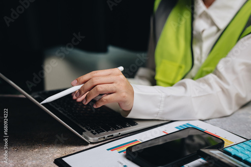 Professional in Safety Vest Engaged in Work with Laptop and Digital Tools in Modern Office Environment