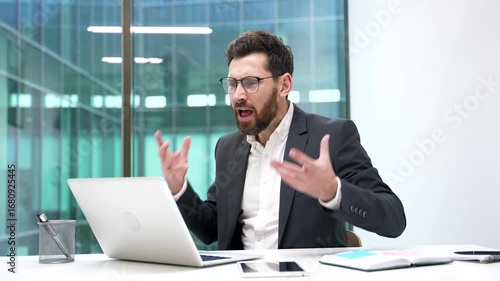 Angry businessman arguing talking on video call using laptop computer sitting at workplace in business office. Excited dissatisfied boss in formal suit scolds subordinate, solving stressful situation.