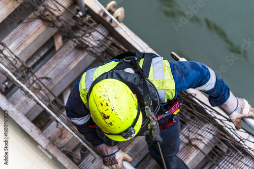 A deckhand prepares the pilot ladder as the ship readies for port entry. Secured in a safety harness, he carefully ensures the setup is ready, focusing on the task in a high-risk environment. 