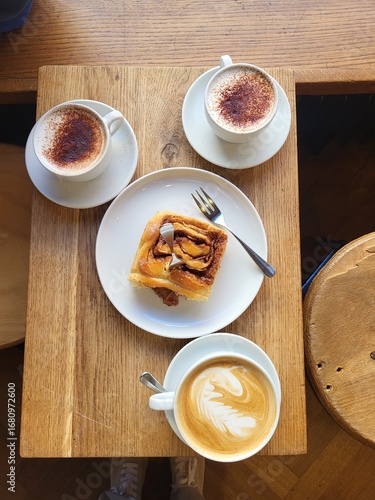 Coffee date with children. 1 cappuccino cup, 1 cinnamon roll, 2 cups of babyccino (milk) are  on wooden rustic table.