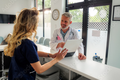 Senior male doctor smiling while showing and explaining information on a tablet to a young female nurse at the hospital reception desk, engaging in a lively discussion about patient care