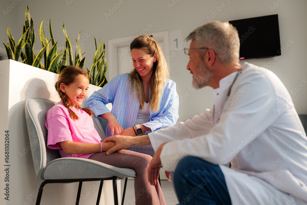Obraz premium Friendly pediatrician talking with a smiling little girl and her mother, reassuring them before a medical visit in a modern clinic waiting room