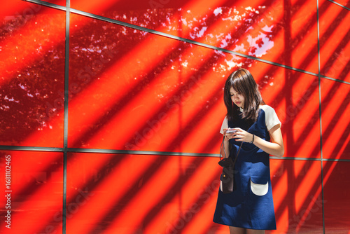 Girl using smart phone while standing near red wall at sunny day