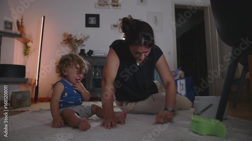 Mother rubbing and cleaning carpet to remove a stain while toddler cries beside her seeking attention, candid indoor family moment showing daily parenting challenges