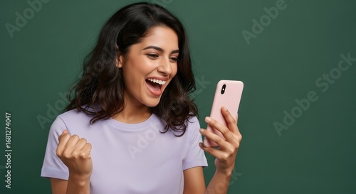 Woman with dark hair and light purple shirt looking at pink phone with excited expression on green background