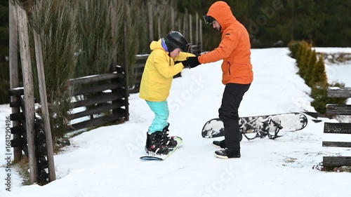 A child receives snowboarding instruction from an adult. They're on a snowy slope near a wooden fence, focusing on balance and technique. The scene evokes winter sports and learning.