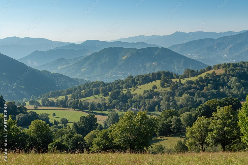 Naklejka premium Tranquil green valley dotted with trees and farmland, surrounded by mist-covered blue mountain ranges, forming a calm and beautiful countryside scene.