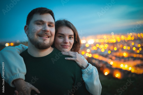 Photos Young family couple embracing on hilltop overlooking city lights at dusk