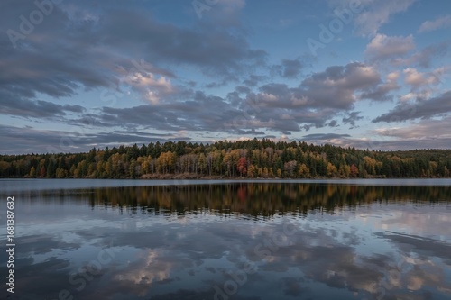 Serene twilight moment over a still lake mirroring the sunset sky, bordered by a forest hinting at autumn hues