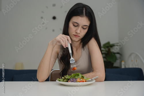 Cuadro en lienzo Young woman looking bored or unhappy while eating salad, pushing food with fork at table