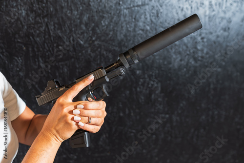 Close-up of a woman's hands with a manicure holding a modern handgun equipped with a silencer and red dot sight.