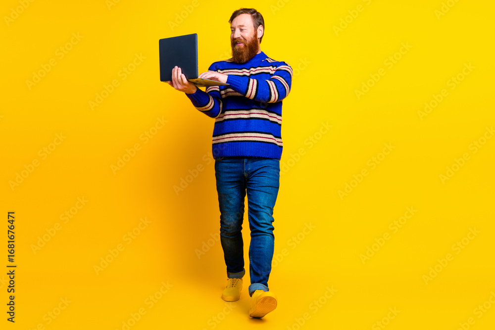 Fototapeta premium Stylish man with a beard holding a laptop while walking against a vibrant yellow background in casual fashion