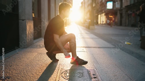 Man crouching on city street at sunrise tying shoelaces in sportswear. Represents preparation, discipline, and determination in active lifestyle and fitness routine
