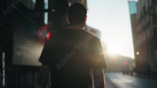 Back view of man walking past modern glass buildings in city street at sunrise. Symbol of reflection, progress, and calm lifestyle in urban environment.
