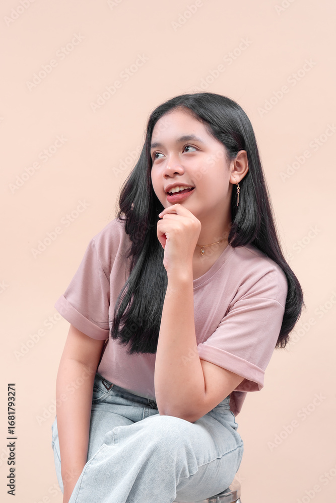 Fototapeta premium Asian woman with long black hair in casual modern outfit, wearing pastel t-shirt and denim, sitting with hand on chin, smiling, showing charm, elegance, and youthful confidence.